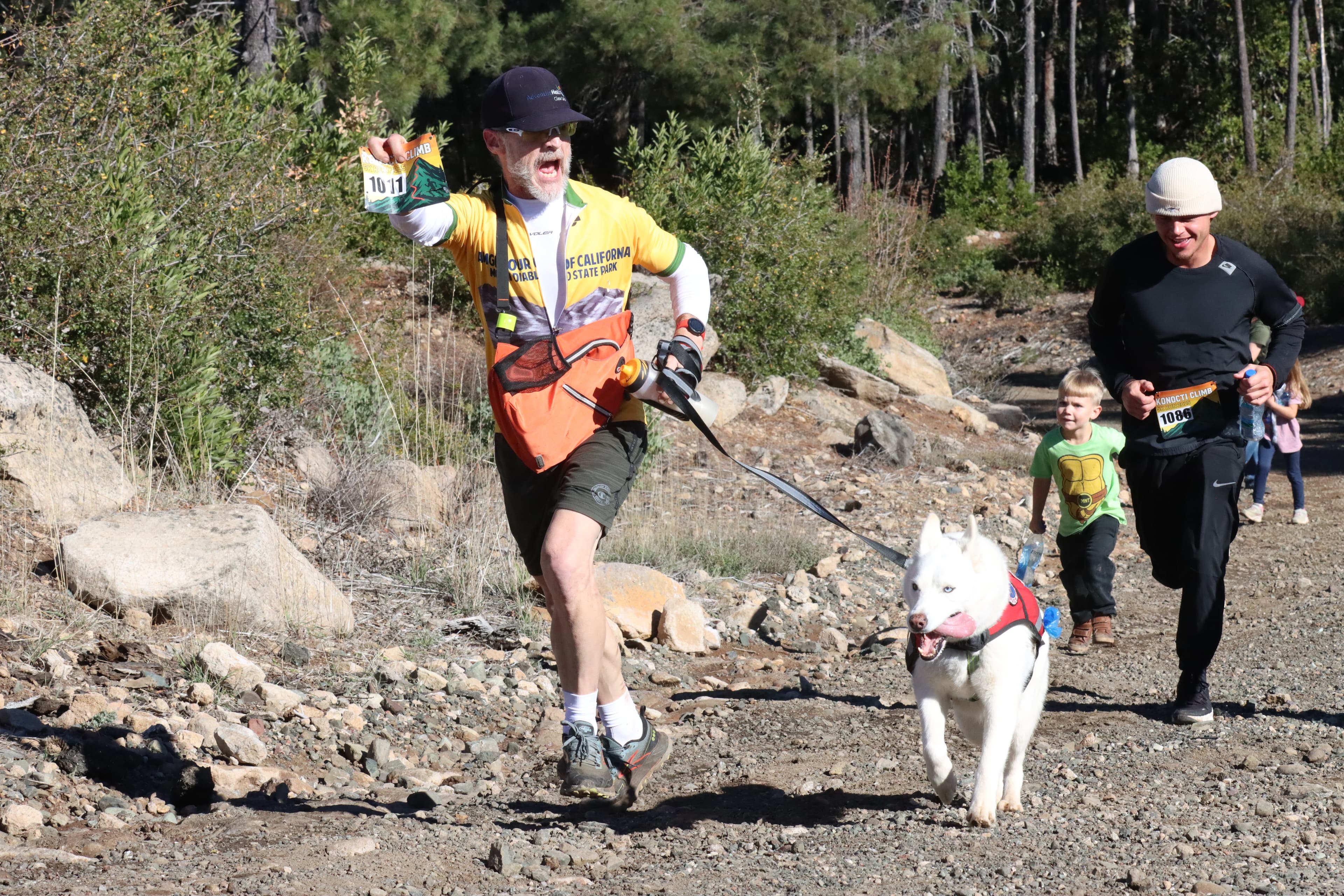 A participant raises a race bib while climbing a rocky trail with a dog and other participants at the 2025 Konocti Climb.
